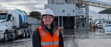 Smiling graduate in a white Holcim hard hat, safety glasses and orange hi-vis vest standing in front of a concrete batching plant with a Holcim agitator truck.