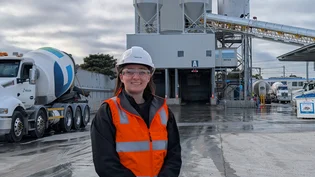 Smiling graduate in a white Holcim hard hat, safety glasses and orange hi-vis vest standing in front of a concrete batching plant with a Holcim agitator truck.