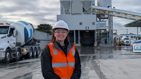 Smiling graduate in a white Holcim hard hat, safety glasses and orange hi-vis vest standing in front of a concrete batching plant with a Holcim agitator truck.