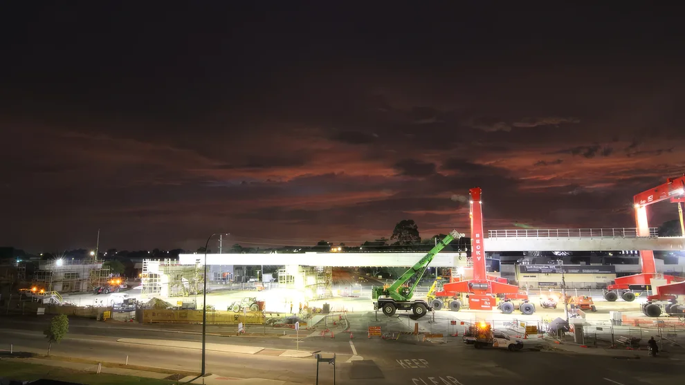 Floodlit construction site at dusk with an elevated rail viaduct spanning a road, red gantry cranes and a mobile crane working on Perth’s Armadale Line