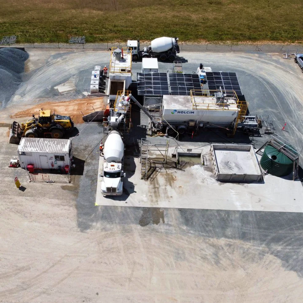 Aerial view of a Holcim mobile concrete plant with rooftop solar panels, a mixer truck and wheel loader on a gravel site.