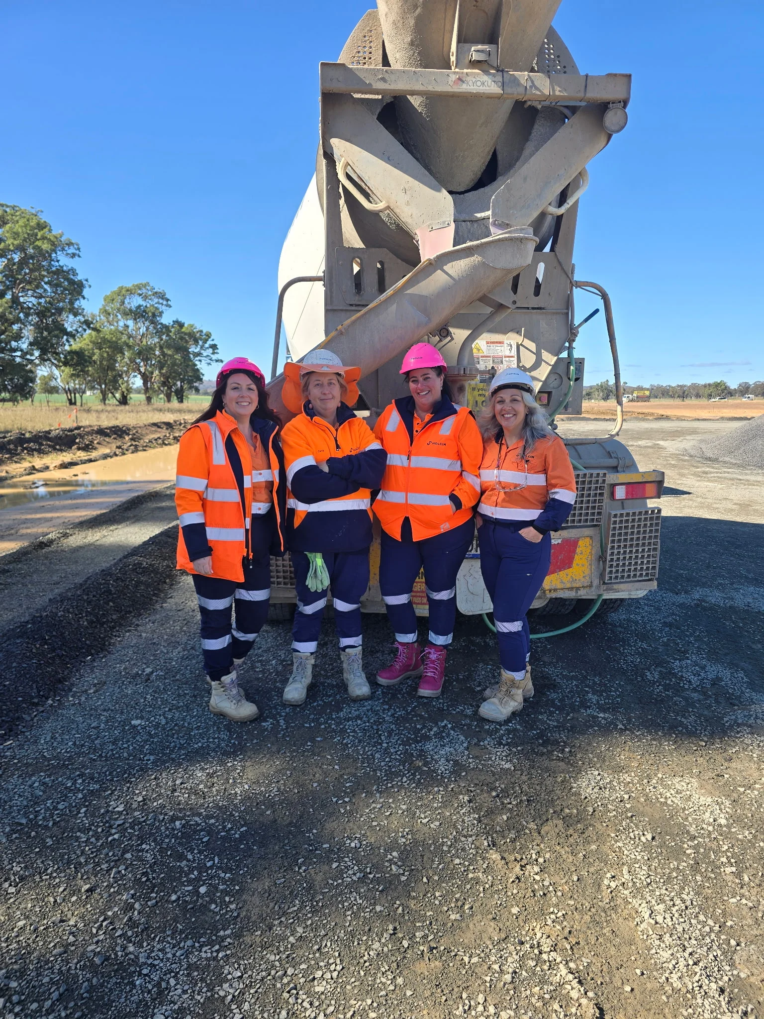 Four women in hi-vis workwear and hard hats stand together in front of a concrete agitator truck on a construction site in Central West NSW.