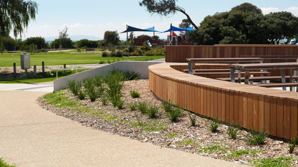 Curved exposed aggregate pathway edging timber seating and native planting at the Rye foreshore, with the bay in the distance.